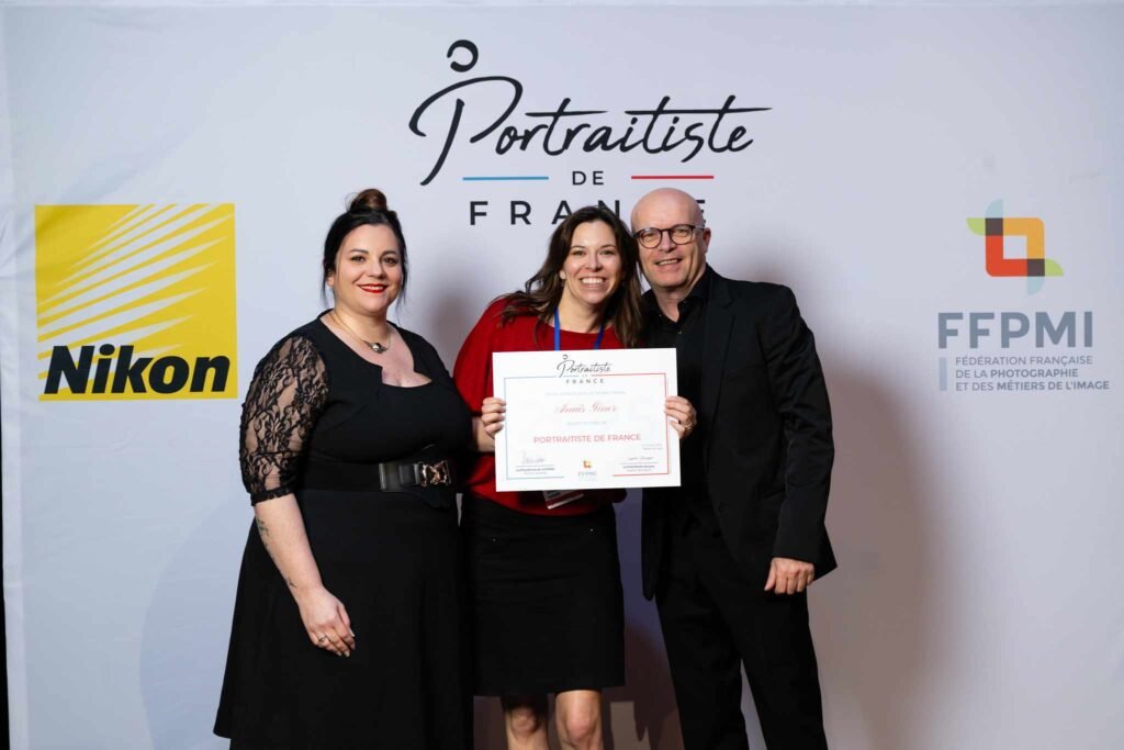 Anaïs Giner tenant son diplôme de Portraitiste de France, entourée de deux personnes devant le fond de la cérémonie Portraitiste de France 2025